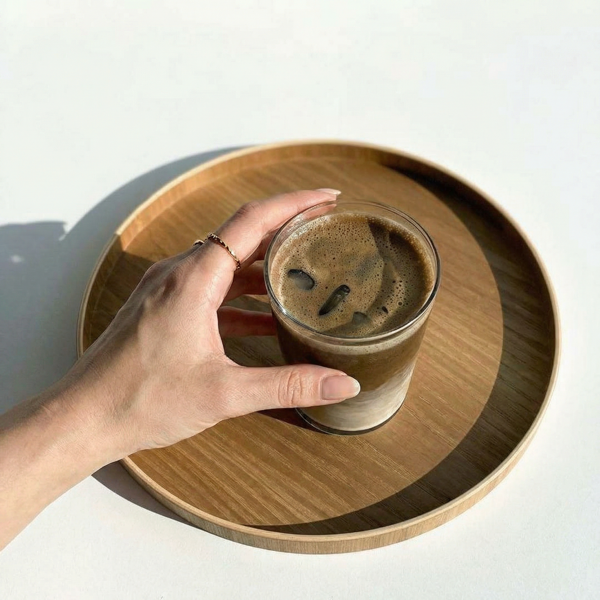 Hand holding a glass of iced coffee on a wooden tray with a white background