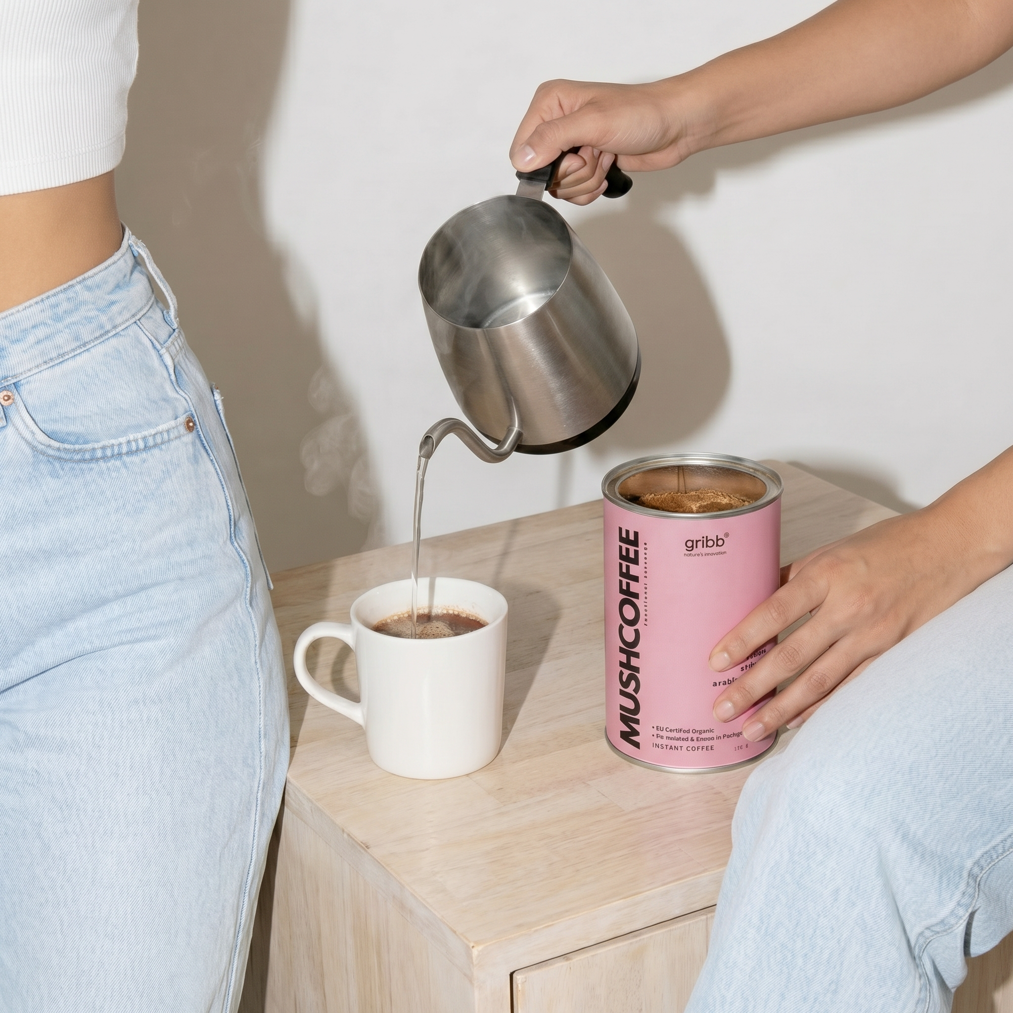 Person pouring coffee from a can into a mug with a pink can labeled 'Mushcoffee' on a wooden surface.