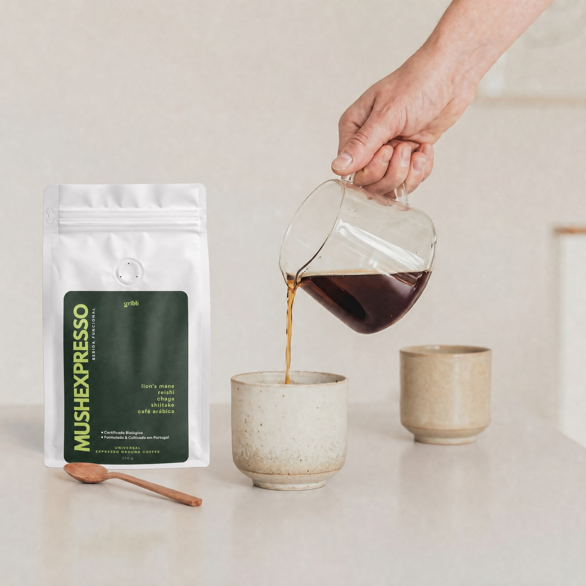 A hand pours freshly brewed espresso from a glass pitcher into a ceramic cup, with a stand-up pouch of Mush Expresso coffee and a small wooden spoon on a light countertop.