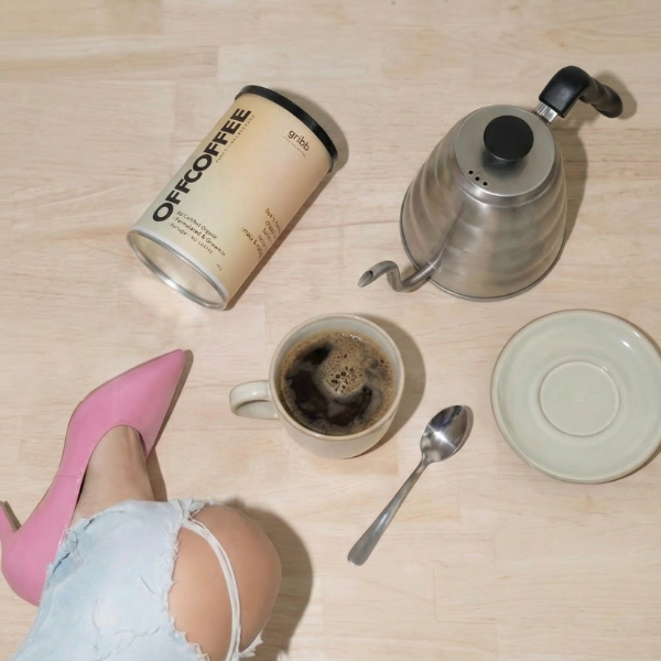 A styled flat-lay of coffee gear on a light wood floor, featuring a can of Off Coffee, a stainless steel kettle, a cup of coffee, a spoon, a saucer, and a person's legs in light blue denim shorts and pink high heels.