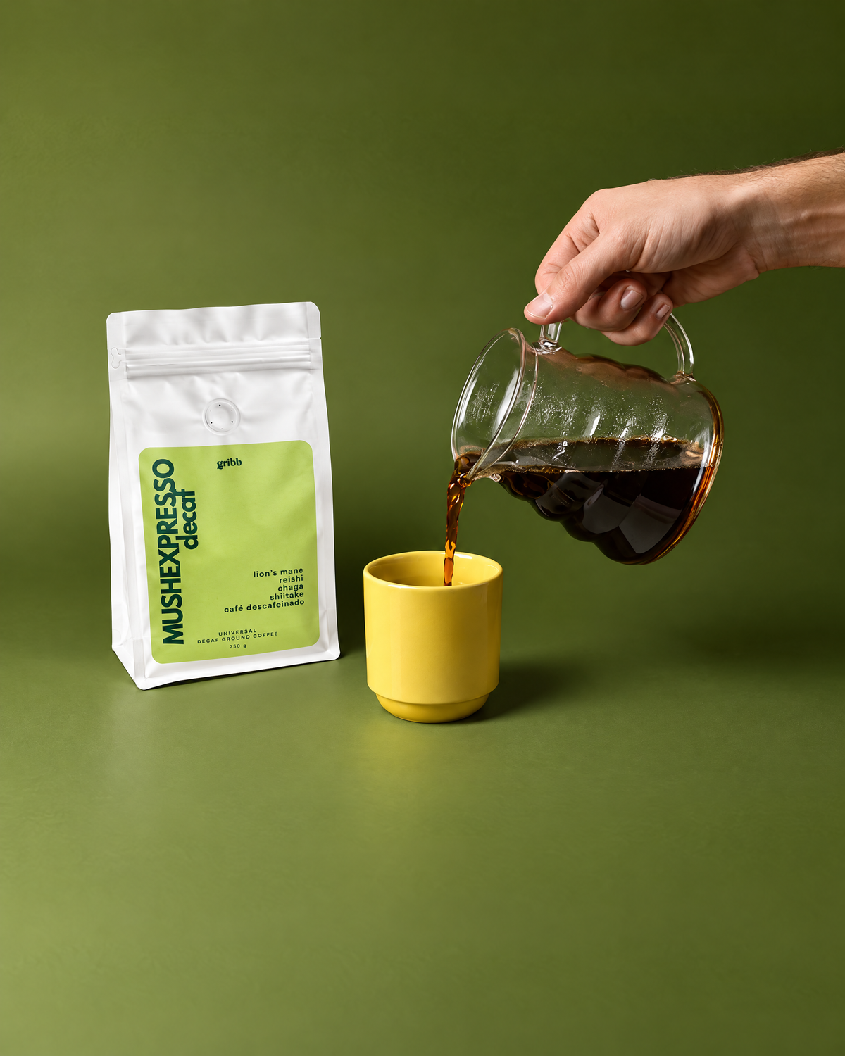 A hand pours dark coffee from a glass pitcher into a yellow cup next to a white bag of mulch espresso roast coffee on a solid olive green background.