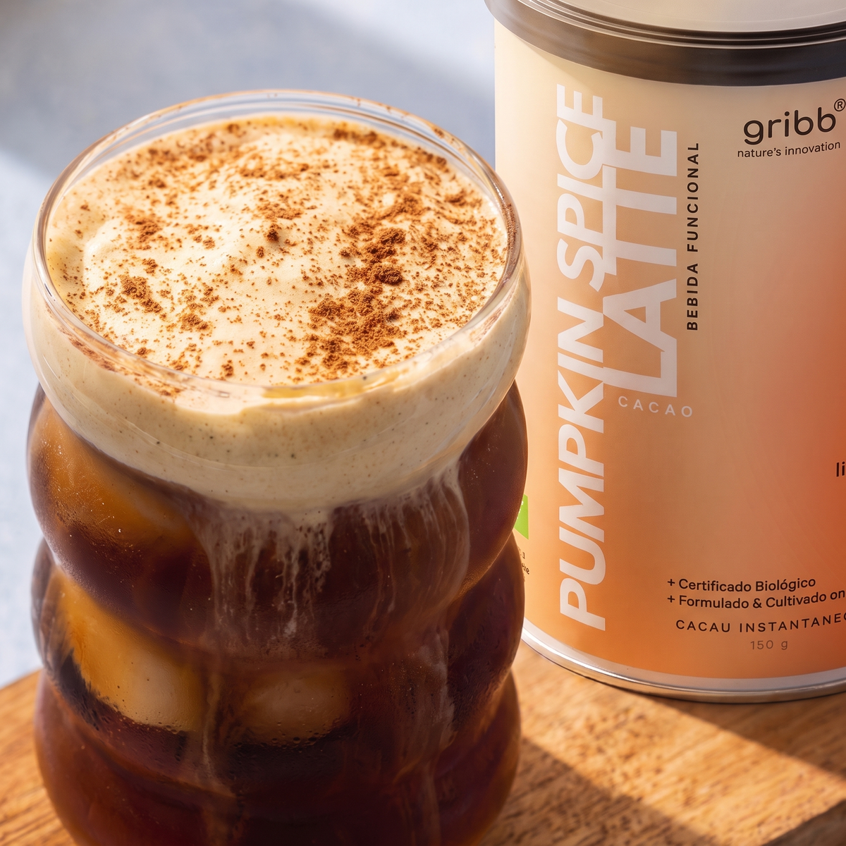 A close-up of a pumpkin spice latte in a clear ribbed glass with whipped cream and cocoa powder on top, next to a can of Gribb pumpkin spice latte powder on a wooden surface.